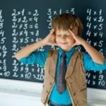 Child stands in front of a chalkboard with his eyes closed and hands to head. The board has multiplication facts written on it.