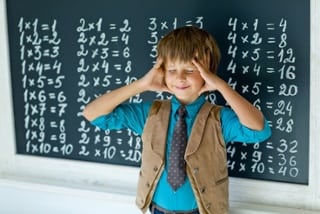Child stands in front of a chalkboard with his eyes closed and hands to head. The board has multiplication facts written on it.